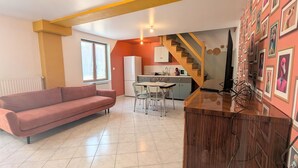 Una sala de estar con sofá, una zona de comedor con sillas, una cocina con refrigerador y una escalera.
