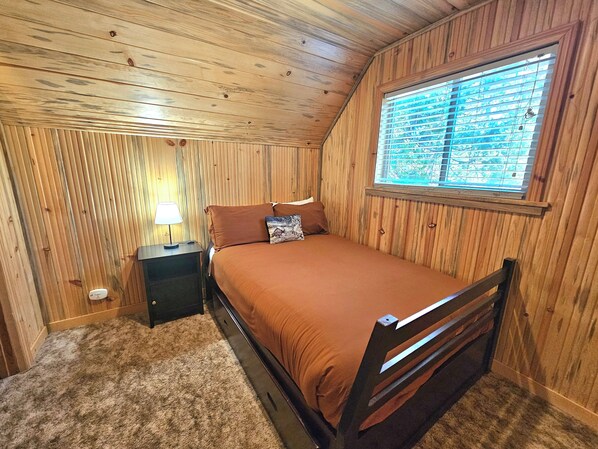 Un dormitorio con techo de madera, una cama individual con ropa de cama marrón, una mesita de noche con lámpara y una ventana con persianas.