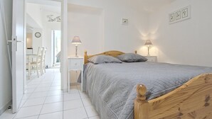 A bedroom with a wooden bed, two bedside tables with lamps, a white door leading to a dining area, and a clock on the wall. - Apartment with terrace at Ars-en-Ré port, free Wi-Fi (Ars-en-Ré)