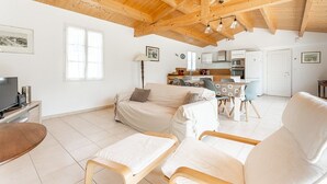 Amplia sala de estar con techo de madera alto, un conjunto de sofás, una mesa de comedor con sillas, una cocina al fondo y un televisor sobre un mueble.