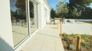 Una casa moderna con un patio que cuenta con una mesa y sillas, una puerta de cristal que da a una terraza, y un jardín con árboles y plantas.