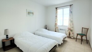 Un dormitorio con dos camas, una silla, una mesita de noche con lámpara, una ventana con cortinas y un cuadro en la pared.
