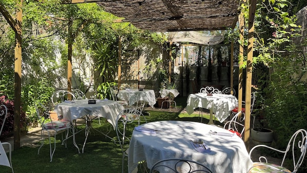 An outdoor patio with tables and chairs, surrounded by greenery and a pergola.