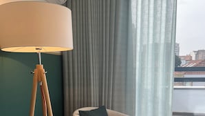 Ein Zimmer mit Stehlampe, Sessel mit Kissen, grauen Vorhängen und Blick auf Gebäude durch ein Fenster.