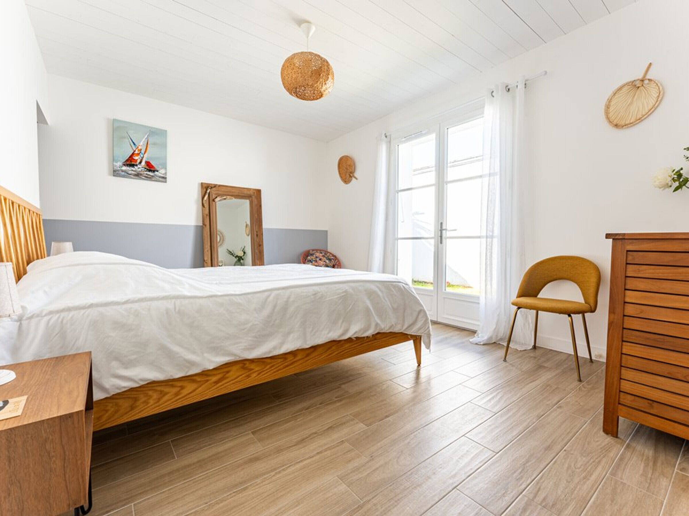 Een slaapkamer met een houten bed, een houten dressoir, een stoel, een spiegel en een raam met gordijnen.