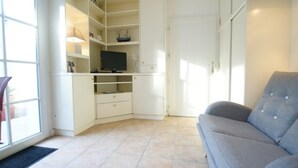 Un salon avec un canapé gris, un téléviseur posé sur une étagère et une porte donnant sur une autre pièce.
