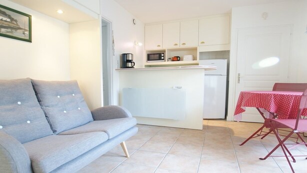 Un espace de vie compact avec un canapé gris, un petit coin cuisine doté de meubles blancs et une table rouge recouverte d’une nappe blanche.