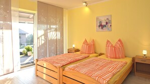 A bedroom with two beds, a sliding glass door, a small bedside table with a lamp, and a painting on the wall.