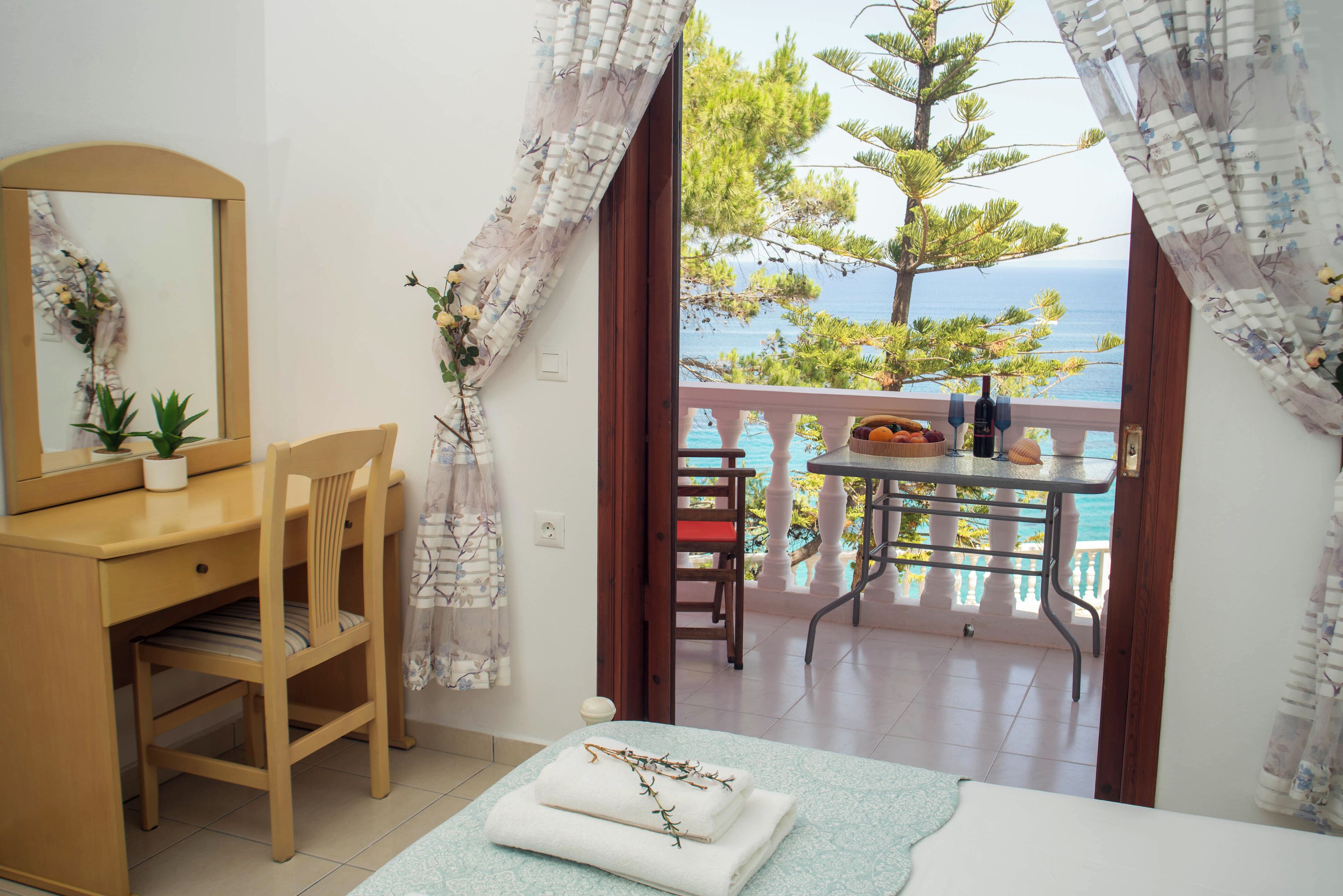 A bedroom with a wooden desk, a chair, a bed with a view of the sea, a balcony with a table and chairs, and a potted plant.