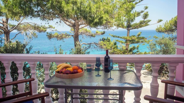 Ein Balkon mit einem für eine Mahlzeit gedeckten Tisch, mit Blick auf eine Küstenlandschaft mit Bäumen und dem Meer.