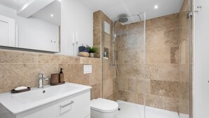 Un baño moderno con cabina de ducha de vidrio, un lavamanos blanco con un soporte de madera para jabón y un inodoro.