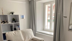 Una sala de estar con un sofá, una mesita redonda, un sillón gris, una estantería y una ventana con cortinas.