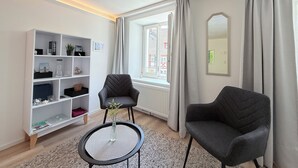 Un salón moderno con una estantería blanca, dos sillones grises, una mesita redonda con un jarrón y una ventana con cortinas.