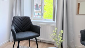 Un salón moderno con un sillón gris, una ventana con vistas a un edificio y vegetación, y un radiador abajo.