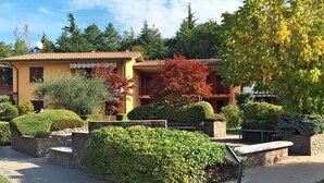 A two-story building with a tiled roof, surrounded by well-maintained hedges and trees. - Beautiful apartment for 3 guests with A/C, pool, TV, balcony and pets allowed (Lazise)
