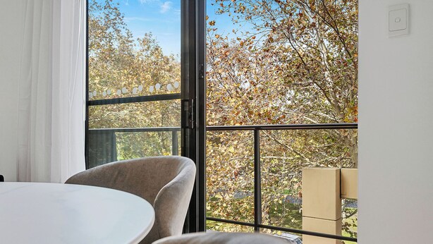 A room with a round table and two chairs, a large window with a view of trees, and a balcony railing. - Urban Escape 2BR Wifi Parking (Perth)