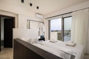 A modern hotel room with a large bed, a wooden bench, a robe rack, and a view of the outdoors through a large window.