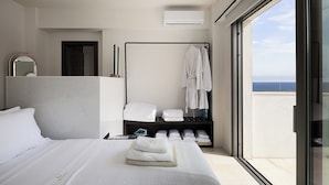 Habitación de hotel con cama, tocador con espejo, perchero y vista al mar a través de un ventanal grande.