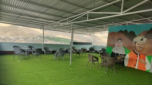 An outdoor seating area with artificial grass and wicker chairs, a mural on the wall, and a view of a distant town. - Universal Comfort Guest House (Kargil)
