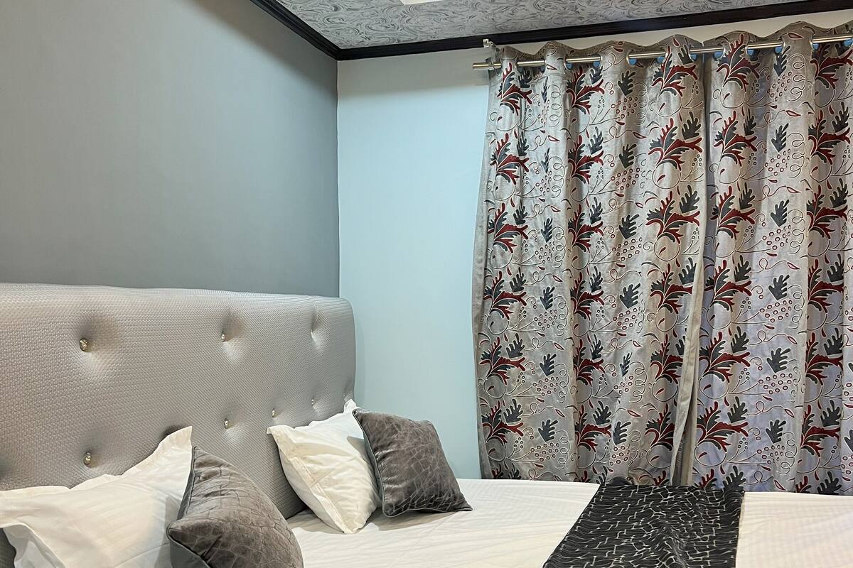 Une chambre à coucher avec un lit, des oreillers, une tête de lit, un rideau et un luminaire au plafond.