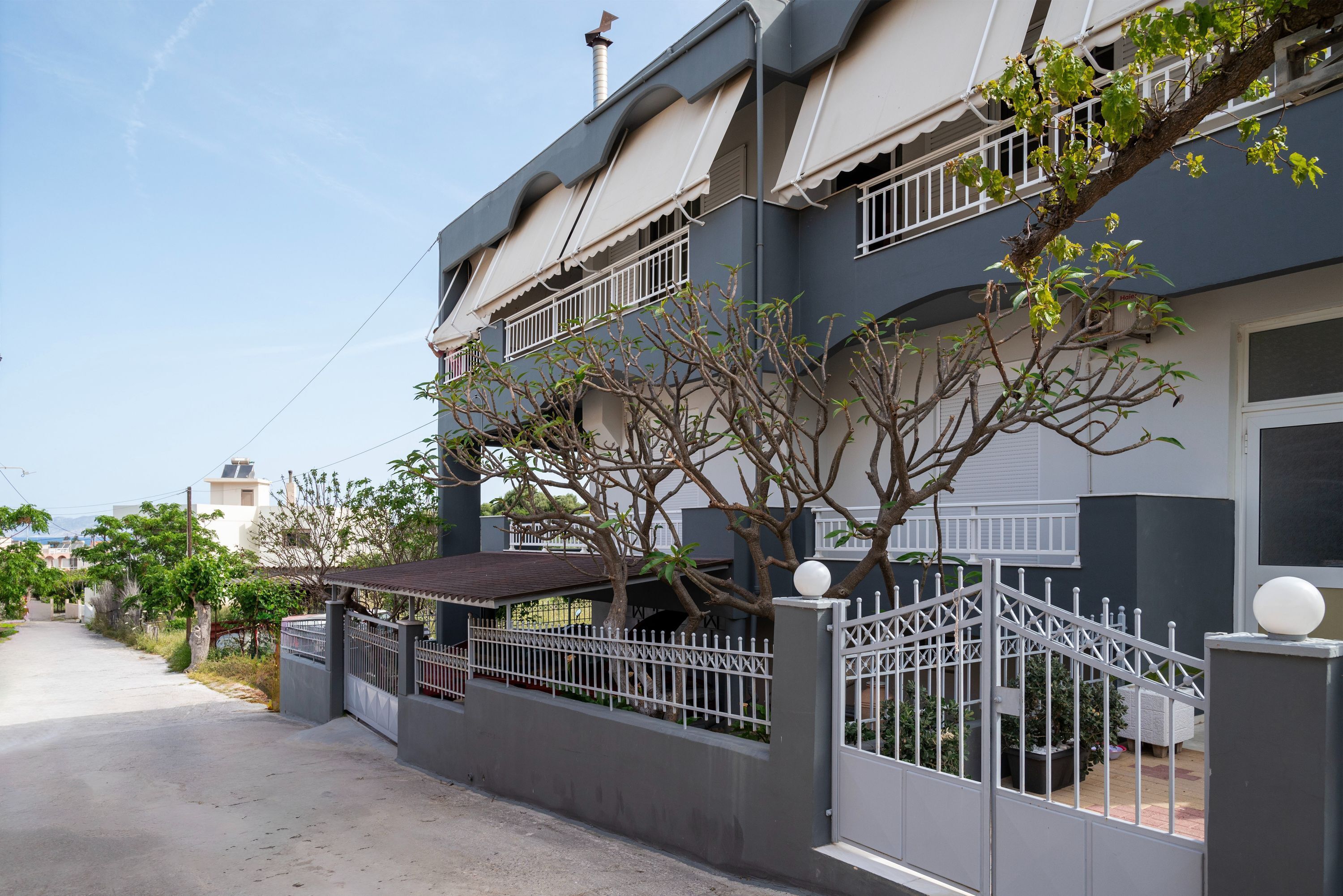 Um edifício residencial de dois andares com fachada cinza, varanda branca e entrada com portão.