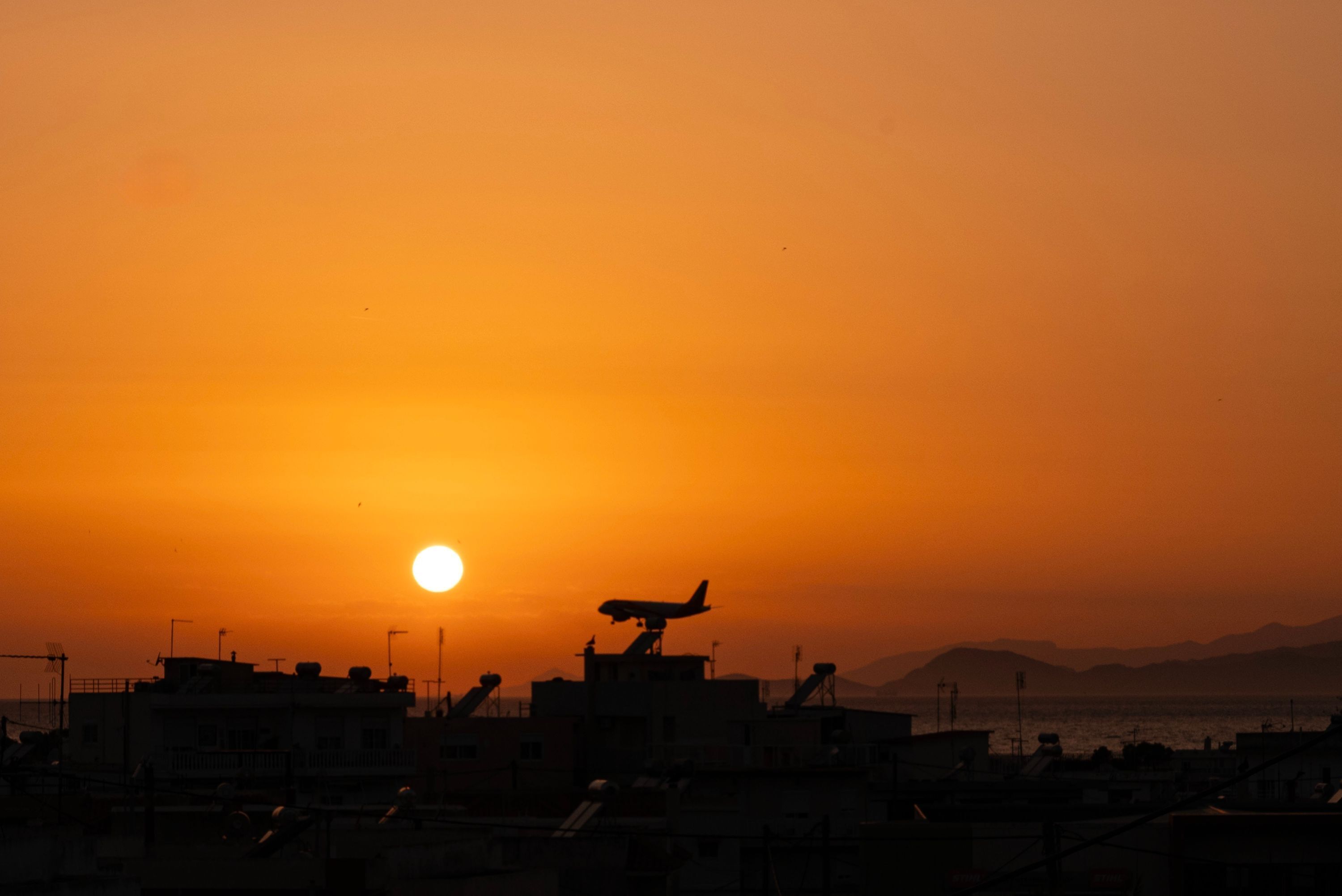 Ein Sonnenuntergang mit einem Flugzeug auf der Startbahn, Gebäuden im Vordergrund und Bergen im Hintergrund.