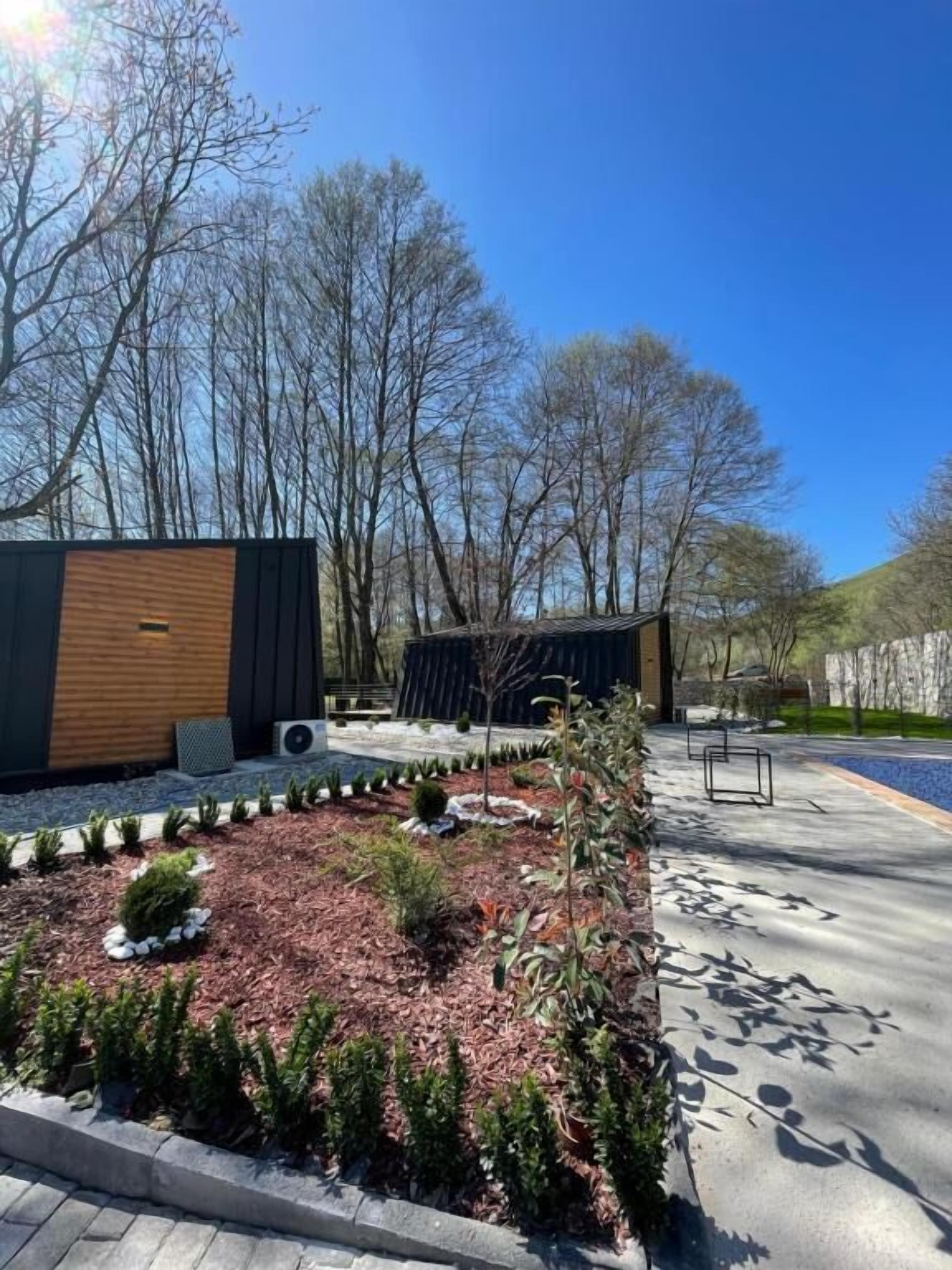 A modern outdoor area with a wooden structure, a paved walkway, and a landscaped garden with small shrubs and a tree.
