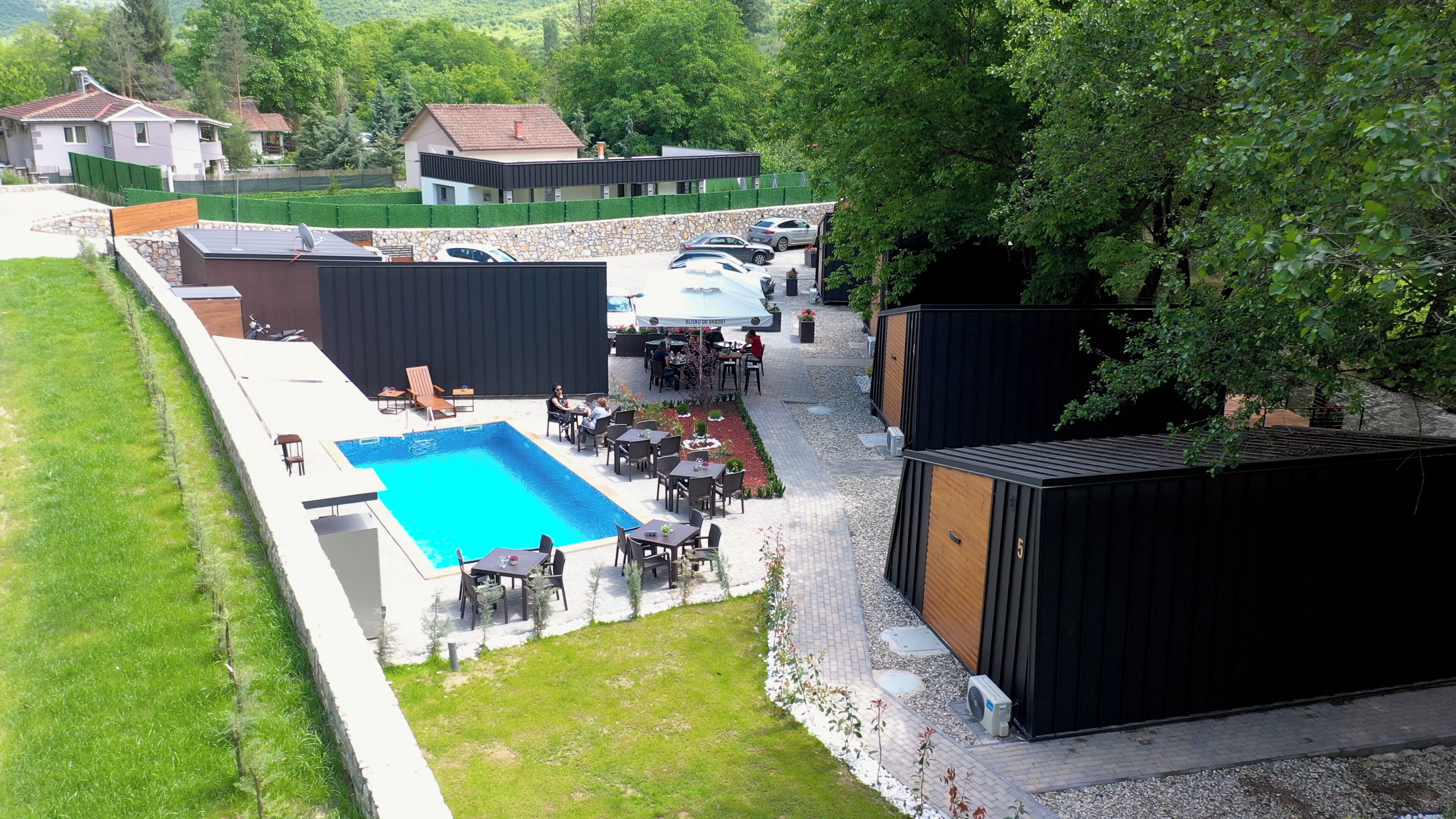 An outdoor pool area with seating and a covered patio, surrounded by greenery and buildings.