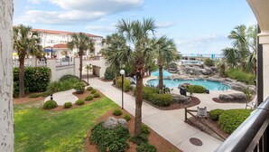 A pool area with palm trees, a stone wall, and a building in the background. - Sunny Side Up at Villa Capriani Resort | Unit 108A | North Topsail Beach, NC (North Topsail Beach)