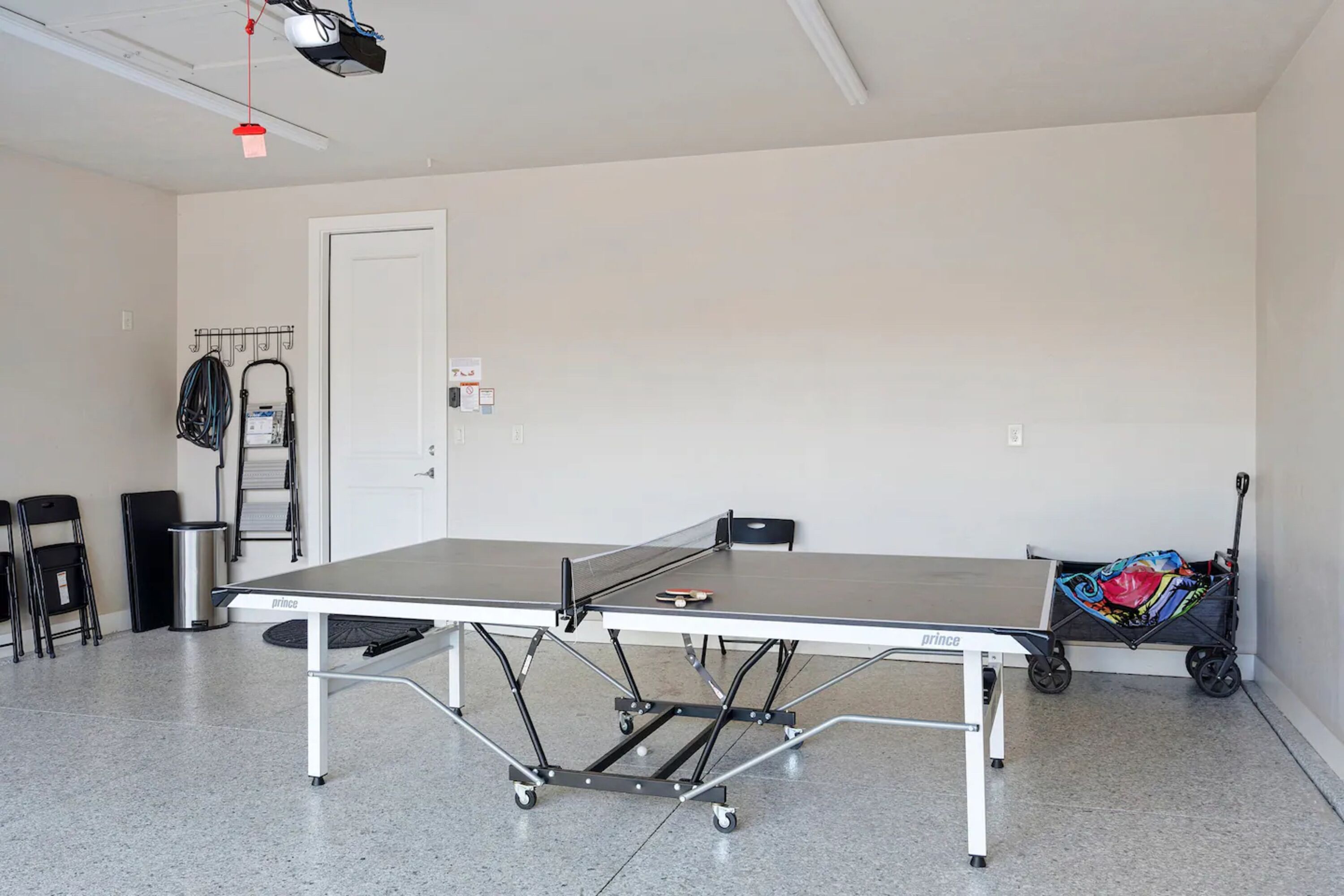 A ping pong table with wheels, a trash can, and a chair in a room with a door and a wall-mounted rack.