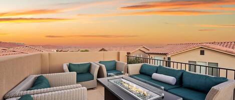 A rooftop patio with wicker furniture and a fire feature, overlooking a cityscape at sunset.