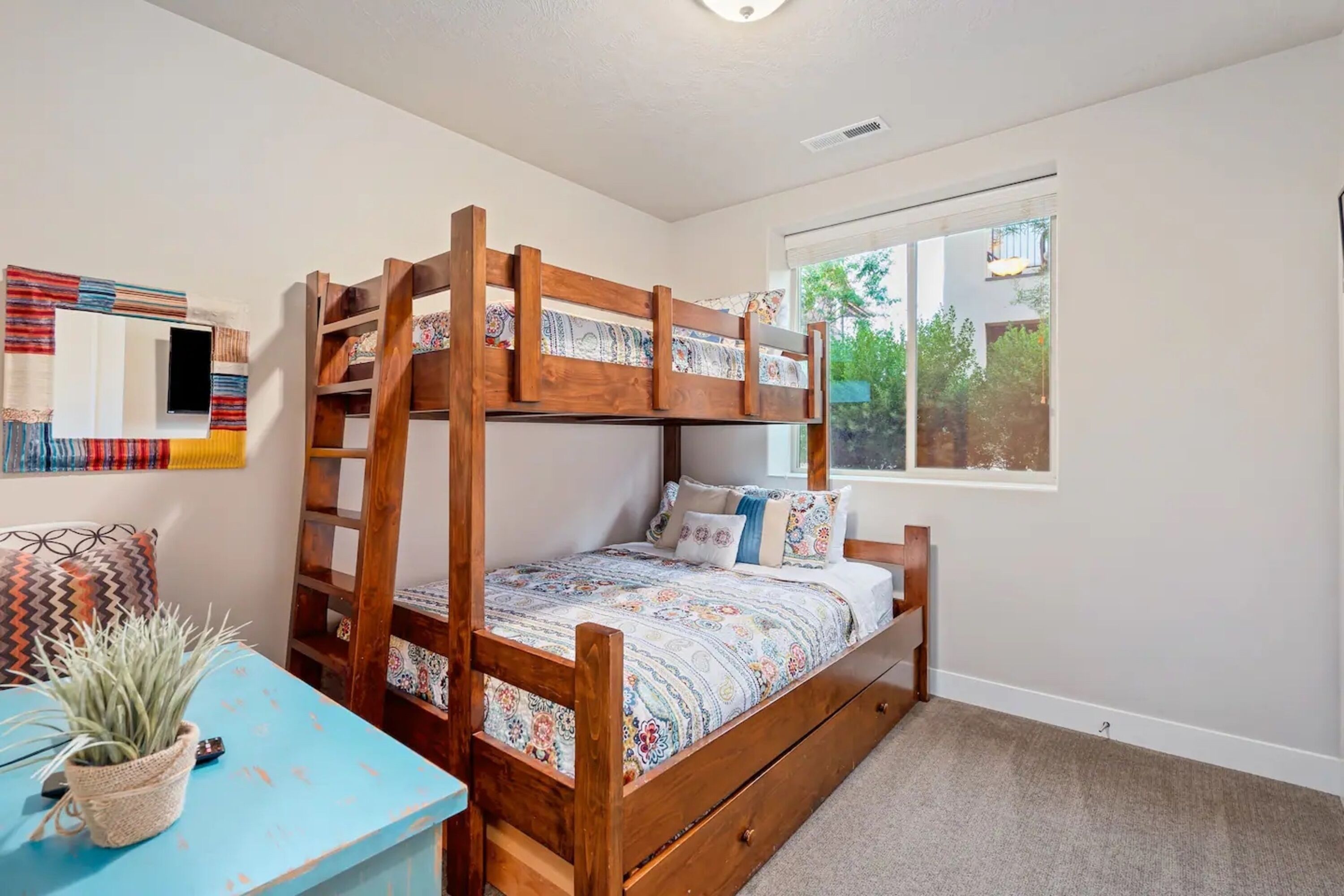 A bunk bed with a patterned bedspread, a small table with a plant, and a window with a view of greenery.