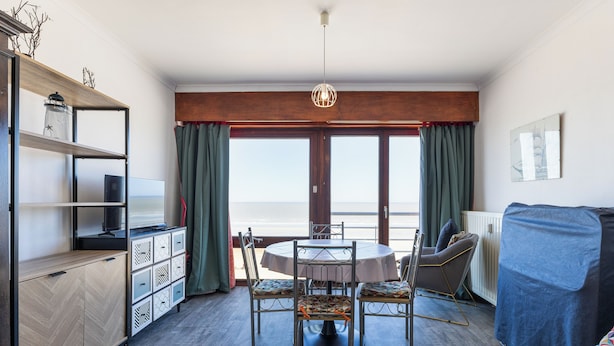 Un comedor con una mesa redonda y sillas, un armario de madera y vista al mar a través de amplios ventanales.