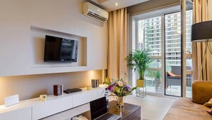 A modern living room with a flat-screen TV mounted on the wall, a coffee table with a laptop and a magazine, a potted plant, and a sofa.