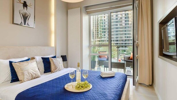 A hotel room with a large bed, a view of a cityscape through a sliding door, and a balcony with outdoor furniture.
