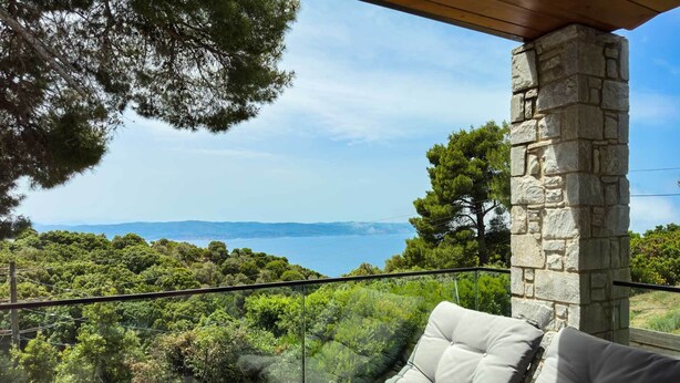 Un balcon agrémenté d’un pilier en pierre, d’un fauteuil rembourré et d’une balustrade en verre, offrant une vue sur un paysage côtier avec des arbres et la mer.