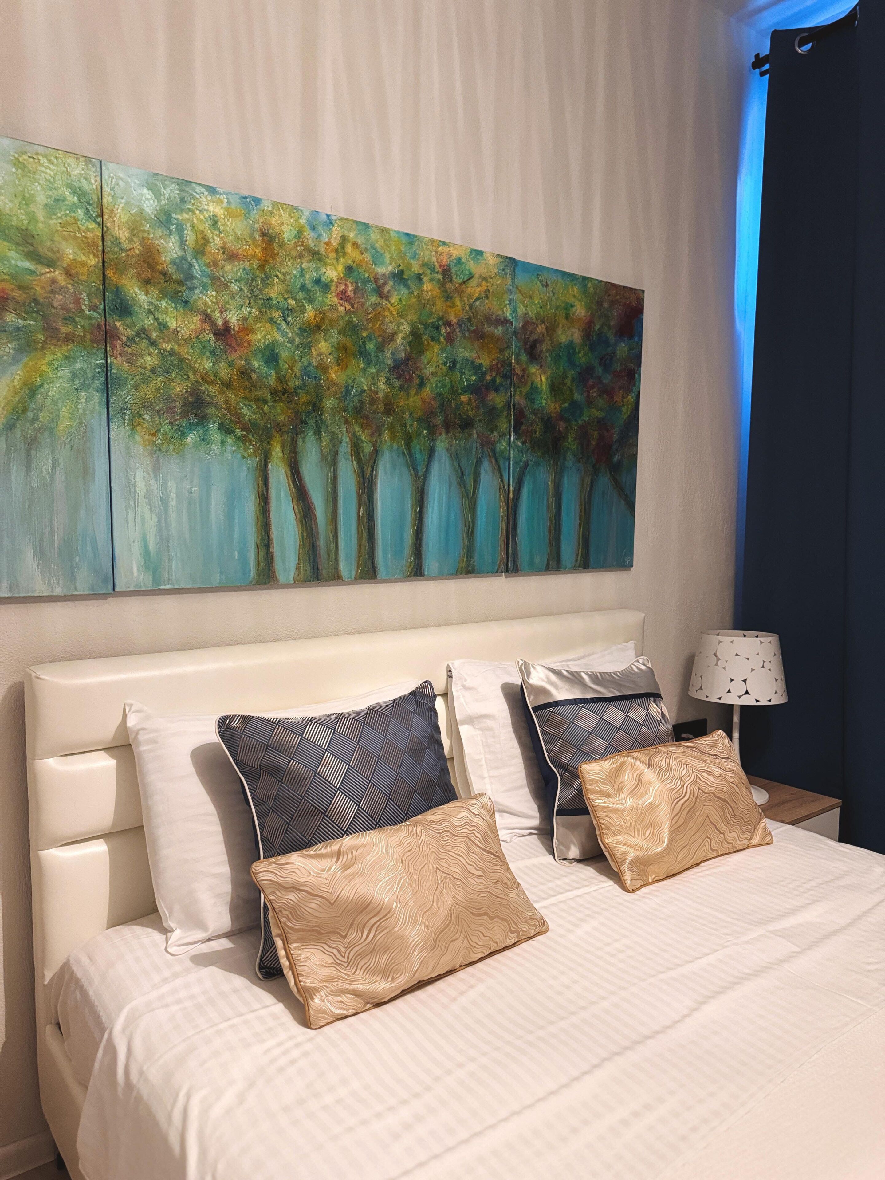 Ein Hotelzimmer mit einem großen Baumgemälde, einem Bett mit vielen Kissen und einem Nachttisch mit einer Lampe.