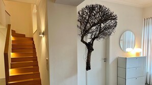 Un interior moderno con una escalera de madera, una escultura de árbol montada en la pared, una puerta blanca y un tocador espejado con luces.