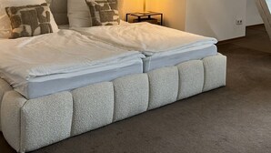 Un dormitorio con una cama grande, dos cuadros enmarcados en la pared, una mesita de noche con una lámpara y un techo inclinado.
