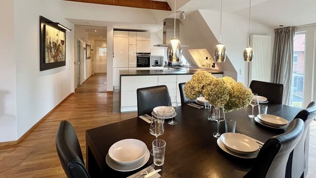 Un comedor moderno con una mesa de madera oscura dispuesta para seis personas, platos blancos y vasos transparentes. Un jarrón con flores blancas está sobre la mesa. La habitación tiene un techo alto con vigas de madera a la vista y se puede ver una cocina y un área de estar al fondo.