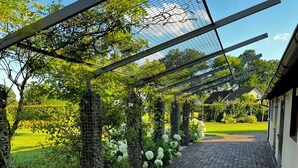 Un sendero de ladrillo que conduce a un área cubierta con techo de metal, rodeada de vegetación y una casa al fondo.