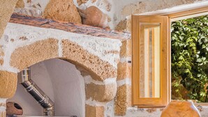 Un interior rústico de piedra con una mesa de madera y sillas de mimbre, un hogar de piedra y una ventana con vistas a la vegetación.