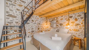 Un dormitorio con pared de piedra, escalera de madera, cama con ropa blanca y lámpara colgante.