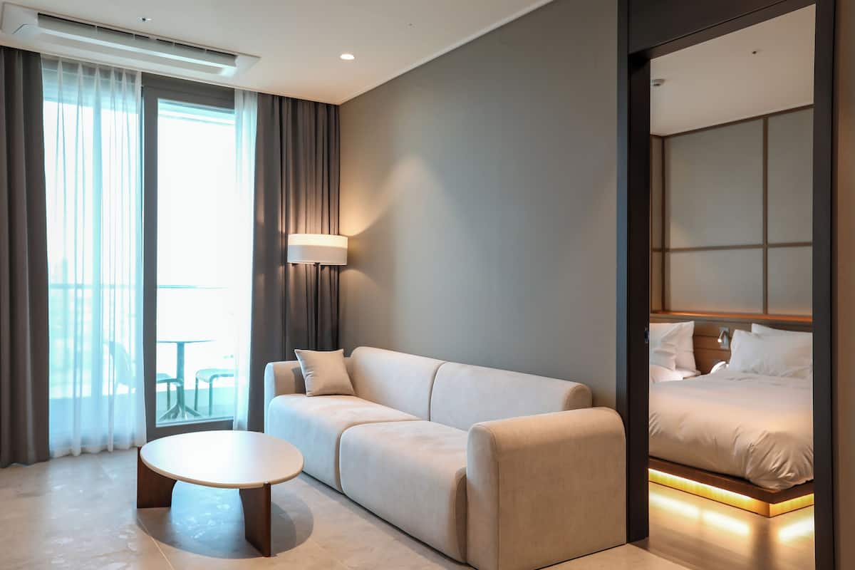 Een moderne hotelkamer met een bank, een rond salontafeltje, een bed met hoofdeinde en uitzicht door een raam met gordijnen.