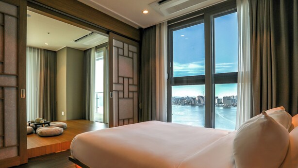 A modern hotel room with a large bed, a view of a cityscape through floor-to-ceiling windows, and a seating area with a table and cushions. - Hotelaround Sokcho (Sokcho)