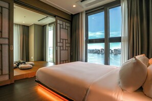 A modern hotel room with a large bed, a view of a cityscape through floor-to-ceiling windows, and a seating area with a table and cushions. - Hotelaround Sokcho (Sokcho)