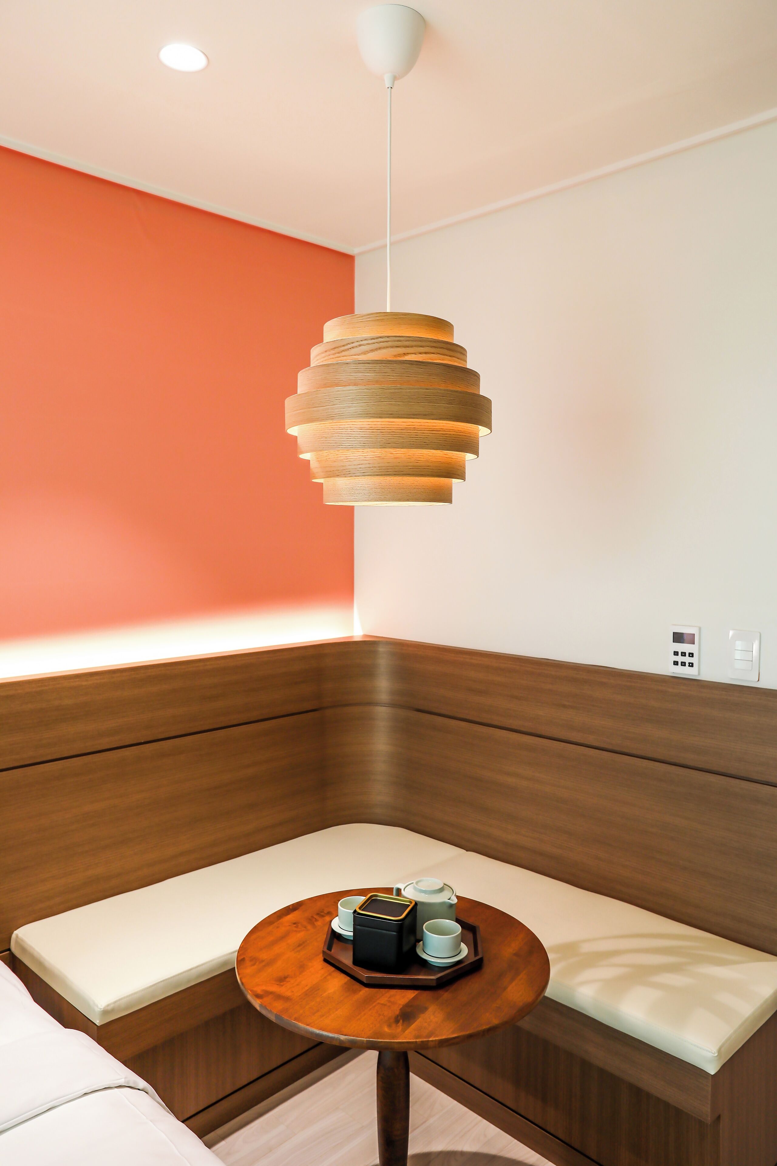 A modern room with a wooden bench, a round wooden table with a tray, a pendant light, and a wall with a red accent.