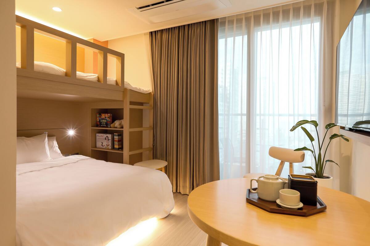 Hotelkamer met een stapelbed, nachtkastje met een dienblad, thee en een kop, een bloempot en een groot raam met gordijnen.