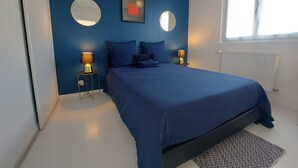 Un dormitorio con una cama grande, dos mesitas de noche con lámparas, un espejo redondo en la pared y una ventana con cortinas.