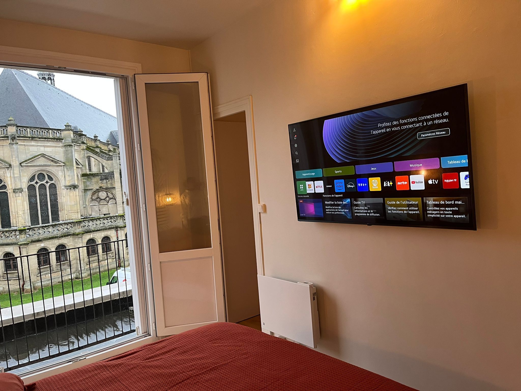 A hotel room with a large window overlooking a historic building, a flat-screen TV mounted on the wall displaying various apps, and a red bedspread.